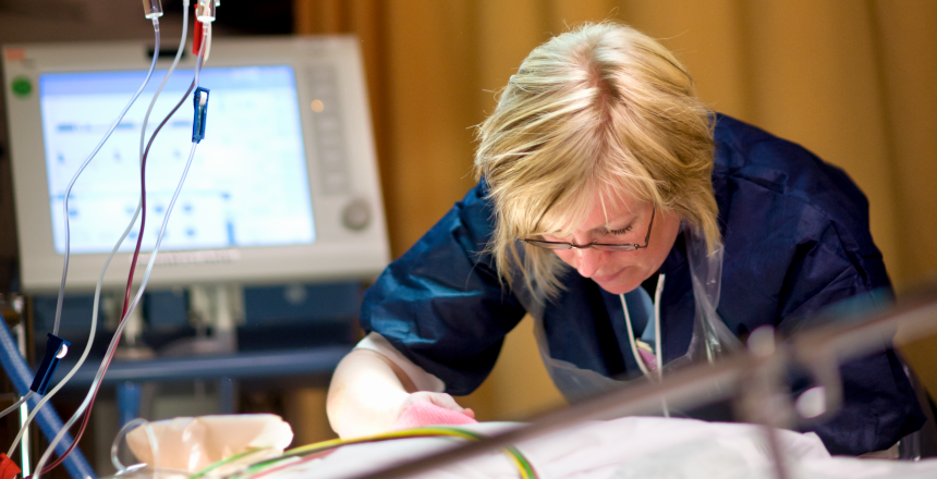 Sjuksköterska böjd över patientsäng, till vänster skymtar droppslangar, i bakgrunden en monitorskärm
