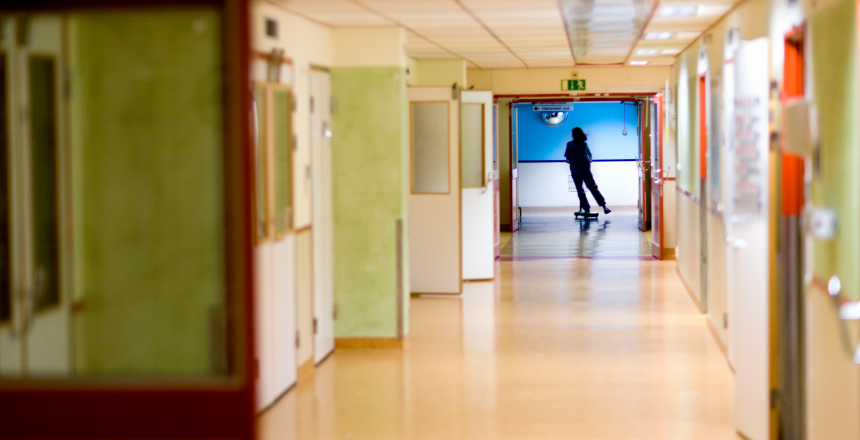 Sjukhuskorridor med öppna dörrar till vänster. I bakgrunden ses en person på sparkcykel.