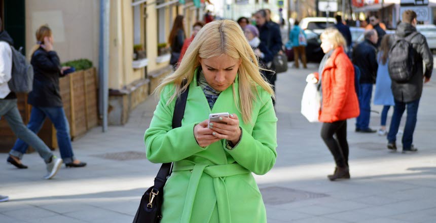 Kvinna i långt blont hår, med illgrön jacka går med mobiltelefonen i handen och stirrar ner i den