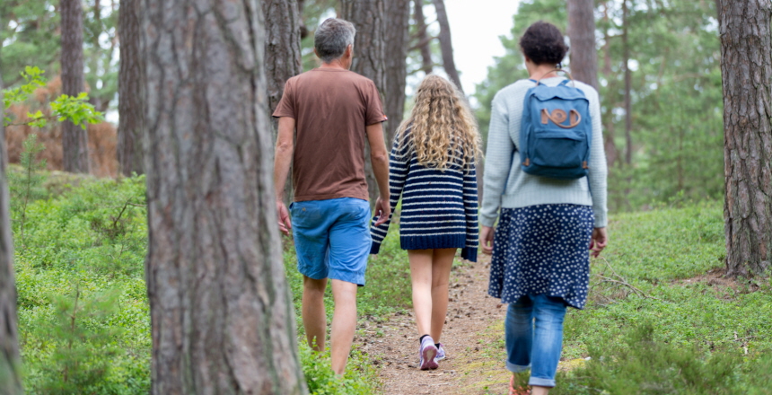 Familj på utflykt i skogen. De promenerar längs en stig. Mamman har en blå ryggsäck på ryggen.
