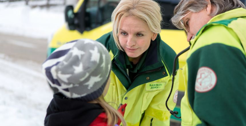 Två sjuksköterskor inom ambulans pratar i förgrunden med ett barn, i bakgrunden ser vi en ambulans