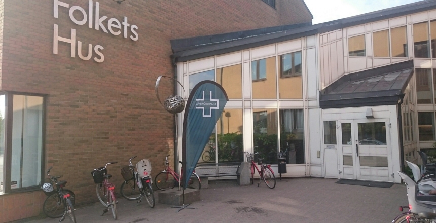 Folkets hus i Halmstad en röd tegelbyggnad med en glasad tillbyggnad. Framför hustet står det cyklar parkerade samt en beachflagga med Vårdförbundets logga på.