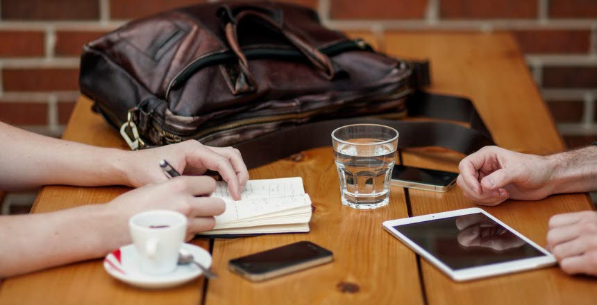 Två personer sitter vid bord och har möte, väska, vatten, glas, kaffe, mobil, telefon, anteckning, penna, hand, händer, skriva, planering, papper, ipad, padda, planer