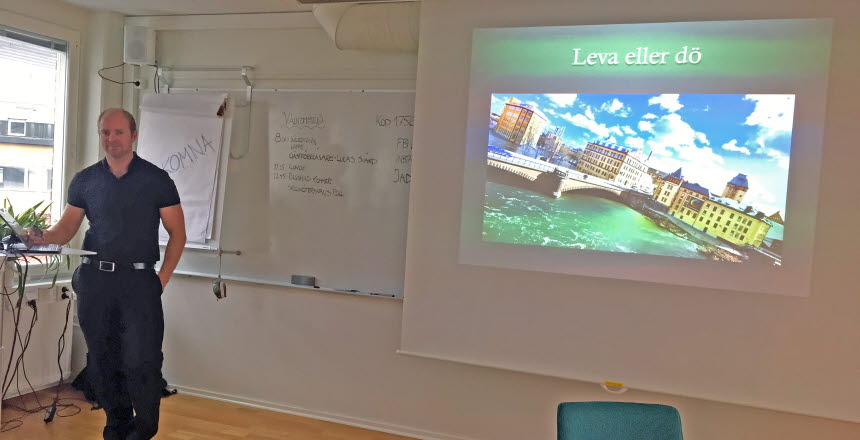 En man i svarta kläder står vid ett bord framme vid en whiteboard och visar en presentation. På filmduken visas en bild av Norrköpings industrilandskap och texten
