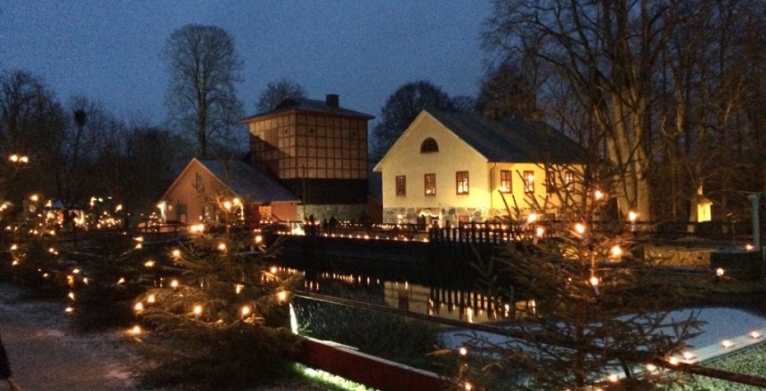 En skymningsbild på adventsgranar med ljus framför en kanal med vatten och en äldre byggnad i bakgrunden.