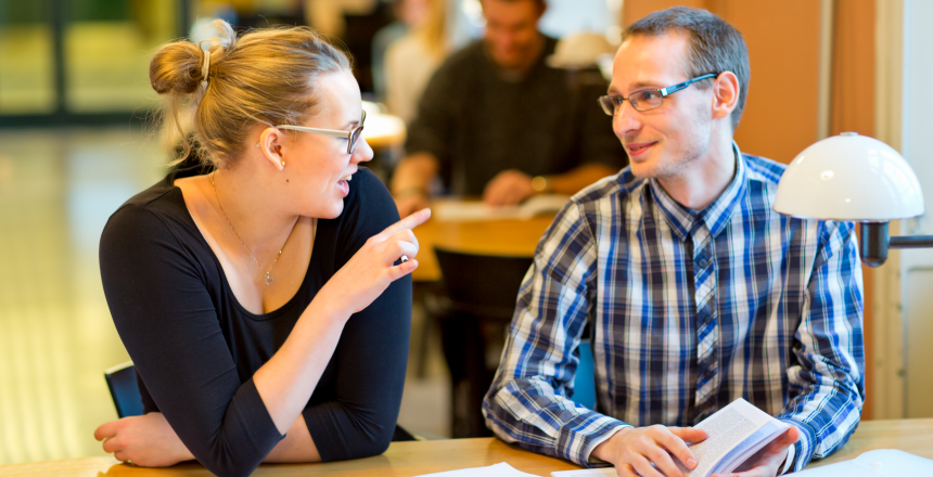 Två studenter, en tjej med uppsatt hår och glasögon och en kille med glasögon och rutig skjorta sitter vid ett bord på ett bibliotek och pratar med varandra.