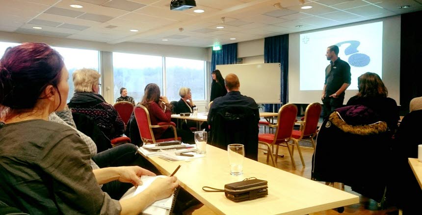 Några personer som sitter i en skolsal och tittar på två personer som står framme vid tavlan och pratar