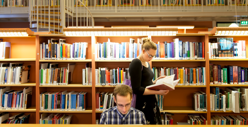 Två studenter på ett bibilotek. Den ena sitter ner och läser, den andra står i bakgrunden och bläddrar i en bok.