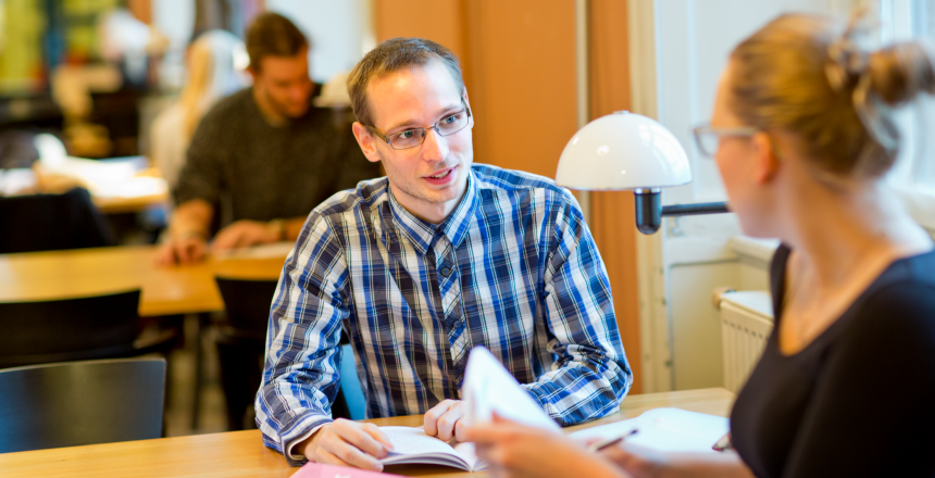 Två studenter, en ung man i rutig skjorta och en ung kvinna med glasögon och uppsatt hår, sitter vid ett bord i ett bibliotek