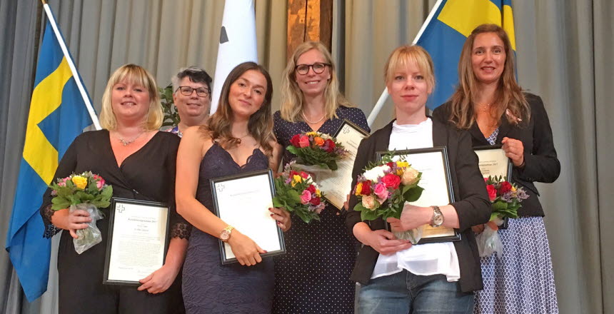Fem uppklädda kvinnor står uppradade framför två svenska flaggor, med diplom och blombuketter i händerna. I bakgrunden står stipendieutdelaren.