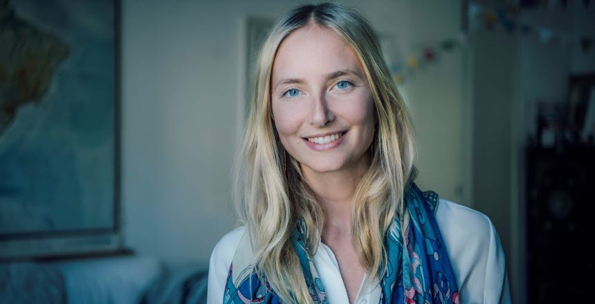 En kvinna tittar leende in i kameran, i bakgrunden syns en tavla på en vägg