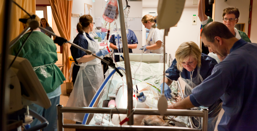 Vårdpersonal står runt en patientsäng där en patient får intensivvård. I förgrunden ses droppställning.