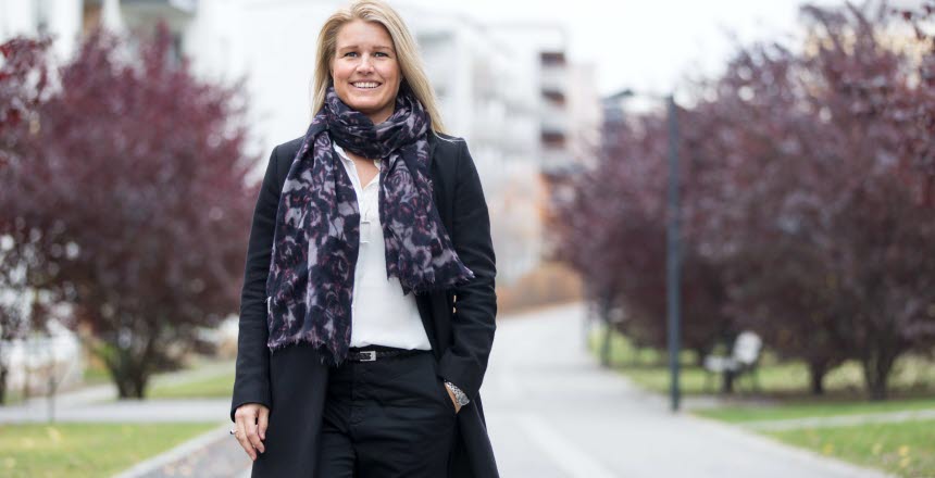 Ljushårig kvinna med svart rock och scarf går leende på en väg kantad av träd