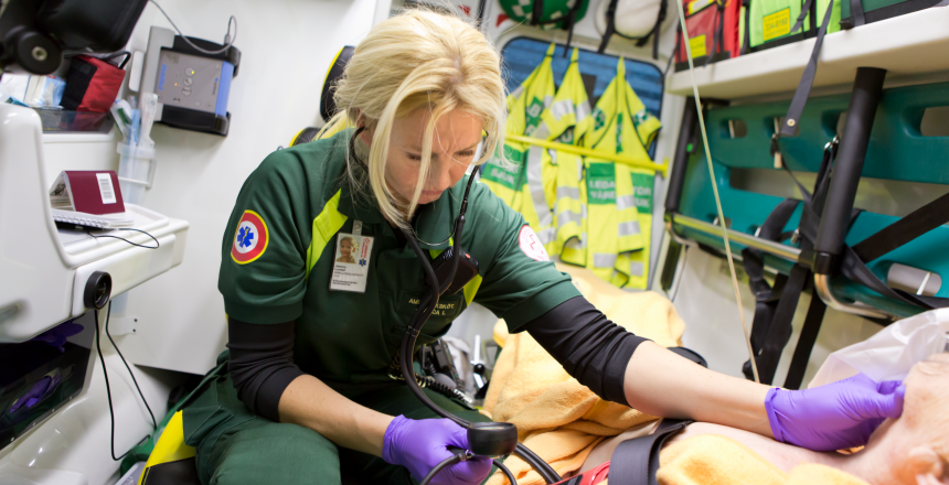 En ljushårig ambulanssjuksköterska mäter pulsen på en patient. Patienten ligger på en bår inne i en ambulans.