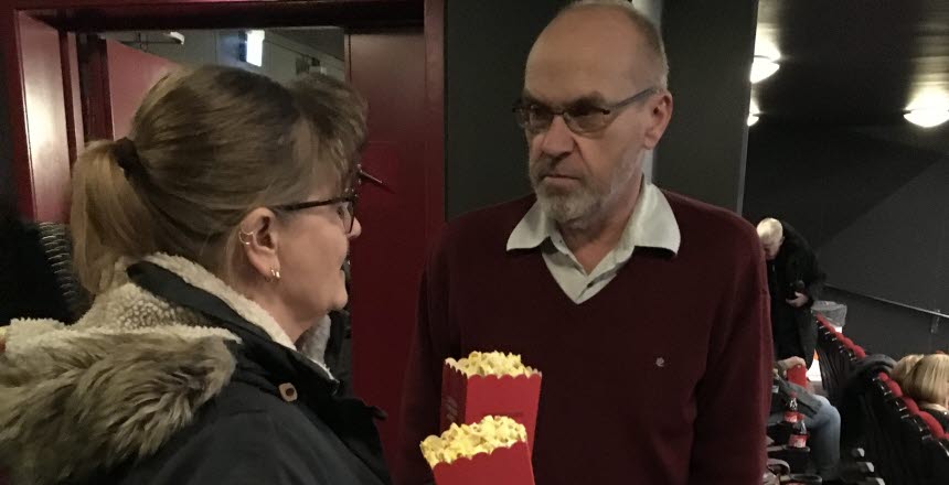 Två personer, en man och en kvinnar står och samtalar längs bak i en biosalong med popcornskartonger i händerna.