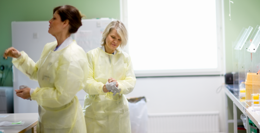 Två kvinnor står i ett laboratorium med ljusa arbetsbänkar på var sida i bilden. Kvinnorna har ljust gula skyddsläder och handskar. En har mörkt uppsatt hår och har vänster sida vänd mot kameran och ryggen åt kollegan. Den andra kvinnan har ljust halvlångt hår och är vänd mot kameran. Hon tar på ett par skyddshandskar. I bakgrunden är det ett stort fönster som släpper in mycket ljus.