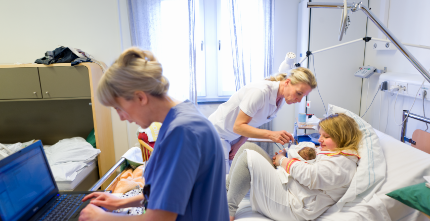 Ett förlossningsrum med mamma och baby till sängs samt en barnmorska vid en laptop och en barnmorska vid sängen